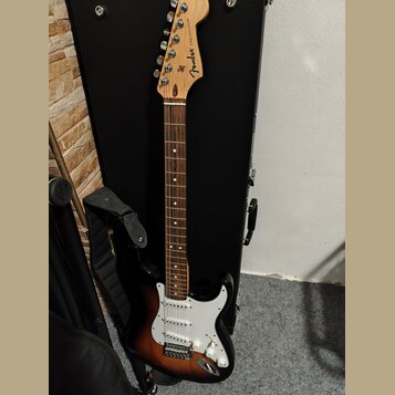 Elektrická kytara s tónem sunburst a bílým pickguardem. Na hlavě hředu je viditelné logo Fender a nápis STRATOCASTER. Kytara má hnědý hmatník a je op…