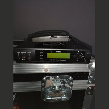 Černý přístroj s nápisem MIPRO a označením M1-808T Stereo Transmitter. Na přední straně jsou umístěny zelený LCD displej, ovládací prvky a konektor p…