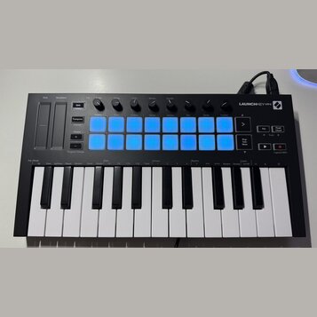 Černý kompaktní MIDI kontrolér s klaviaturou a názvem LAUNCHKEY MINI. Horní panel obsahuje 8 modrých podsvícených čtvercových políček, několik otočný…