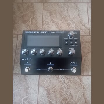 Černý kytarový procesor s nápisem BOSS GT-1000CORE GUITAR EFFECTS PROCESSOR. Zařízení má displej, pět ovládacích knoflíků s čísly 1 až 5, tlačítka EF…