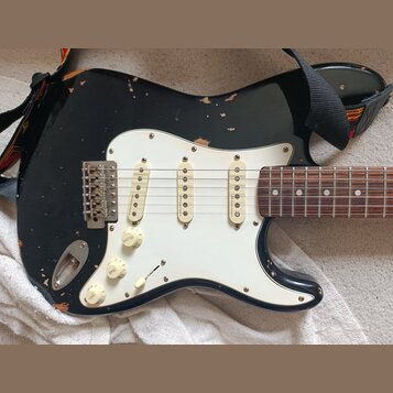 Černá elektrická kytara typu Stratocaster s bílým pickguardem a hnědým hmatníkem. Na každém ze tří snímačů je nápis "Made in USA". Lak na těle kytary…