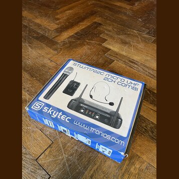 Krabice s bezdrátovým mikrofonním systémem značky skytec. Na obalu je nápis STWM722C MICRO UHF 2CH COMBI a adresa www.tronios.com. Fotografie na krab…