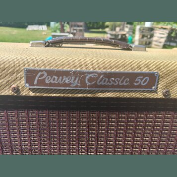 Detail kytarového komba potaženého béžovou texturou s hnědým koženým úchem. Na přední straně je kovový štítek s nápisem Peavey Classic 50. Povrch ští…