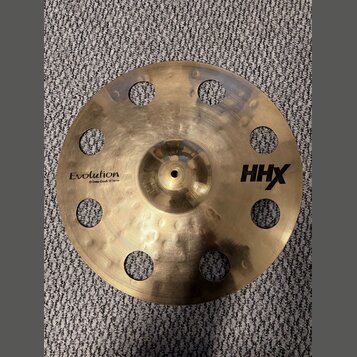 Zlatý kovový činel s otvory v okruhu kolem středu. Na povrchu jsou nápisy Evolution, O-Zone Crash 15" 40 cm a HHX. Povrch vykazuje jemné rýhy a drobn…
