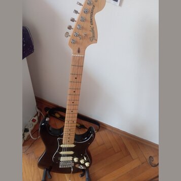 Černá elektrická kytara typu Stratocaster s jasným dřevěným hředníkem a krčkem. Na hlavě je vidět nápis Fender STRATOCASTER. Kytara má tři snímače, b…