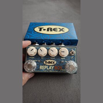 Kytarový efekt s nápisem T-REX REPLAY BOX Delay a doplňkem Made by T-REX Denmark. Zařízení má šedý povrch, čtyři bílé ovládací knoflíky s popisky VOL…