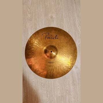 Zlatavý kovový činel s kovaným povrchem. Na horní části je černý nápis Paiste a ve spodní části text Dimensions Hand Hammered Ride. Povrch vykazuje d…
