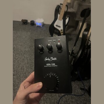 Černý kytarový výkonový zesilovač s nápisem Harley Benton GPA-100 Guitar Power Amplifier. Na horní straně jsou tři menší otočné ovladače s popisky BA…
