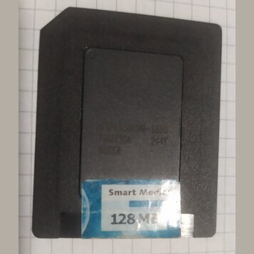 Černá paměťová karta SmartMedia s modrobílou nálepkou. Na nálepce je nápis Smart Media a 128 MB. V horní části těla jsou vyražená písma a čísla: 70NS…