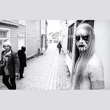 Černobílá fotografie zachycuje muže s dlouhými světlými vlasy a výrazným černým lízením obličeje v ulici s dlážděním. Muž je bez košile a na pravém r…