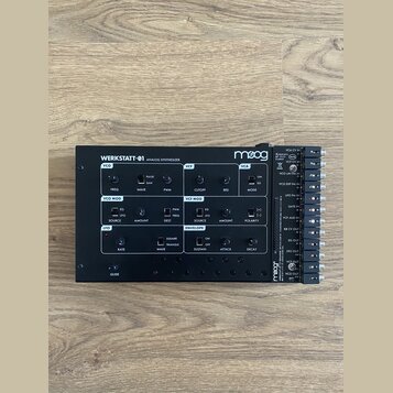 Černý analogový syntetizátor s nápisem WERKSTATT-01 ANALOG SYNTHESIZER a logem moog. Přístroj obsahuje různé ovládací knofíky, přepínače a sekce ozna…