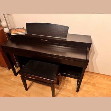 Tmavě hnědý digitální piano v dřevěném provedení s přívodním stolem na noty. Na přední straně je bílý nápis YAMAHA. Součástí je ladící stolek s černý…