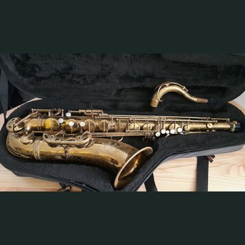 Zlatý saxofon v černém pouzdře. Povrch nástroje vykazuje výrazné opotřebení, škrábance a oděrky laku. Na instrumentu nejsou viditelné žádné značky an…