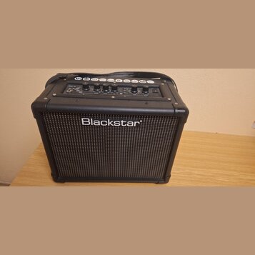 Černý kytarový kombo zesilovač s mřížkou reproduktoru v přední části. Na mřížce je bílý nápis Blackstar. Horní ovládací panel obsahuje několik potenc…