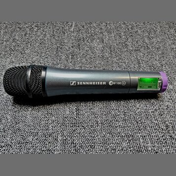 Šedý bezdrátový mikrofon s černou mřížkou a fialovým prstencem u spodní části. Na těle je bílý nápis SENNHEISER s logem a označení ew 100 G2. V zadní…