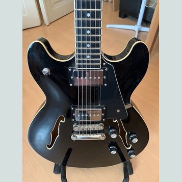 Černá polodublová elektrická kytara s bílým lemováním hran. Nástroj má dva humbuckery, černý plech (pickguard) a čtyři ovládací knoflíky. Na těle jso…