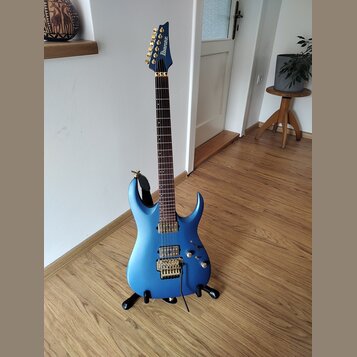 Modrá elektrická kytara s kovovými zlatými komponenty, umístěná na černém stojanu. Na hlavě je viditelné logo Ibanez. Kytara má dva humbuckery, tremo…