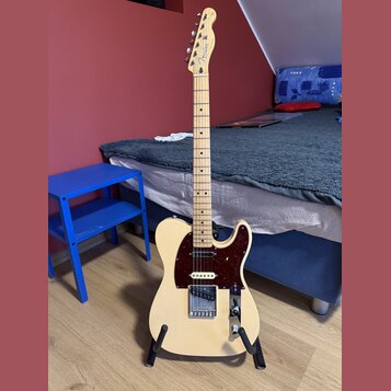 Světle béžová elektrická kytara s tmavě hnědým pickguardem, umístěná na černém stojanu. Na hlavě nástroje je viditelné logo Fender. Kytara má šest st…