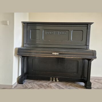 Černý piano v přímém záběru zepředu. Nástroj má zdobný ornament uprostřed klaviaturního víka a tři mosazné pedály. Povrch vykazuje známky opotřebení…