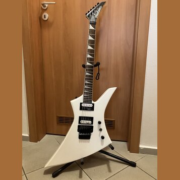 Bílá elektrická kytara s ostře hranatým tvarem těla a špičatou hlavou. Na hlavě je viditelné logo Jackson. Kytara má dva černé humbuckery, dvě ovláda…