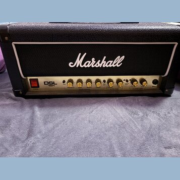 Kytarový zesilovač značky Marshall, model DSL 15H. Má černý texturovaný povrch a bílý lem kolem černé mřížky. Na předním ovládacím panelu jsou umístě…