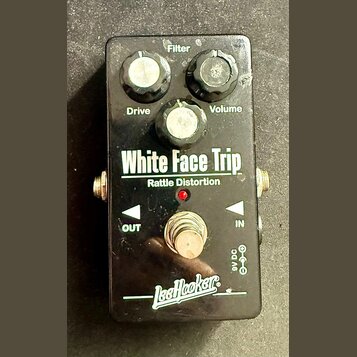 Černý kytarový efekt s nápisy White Face Trip a Rattle Distortion. Na horní straně jsou tři černé otočné ovladače s popisky Filter, Drive a Volume. V…