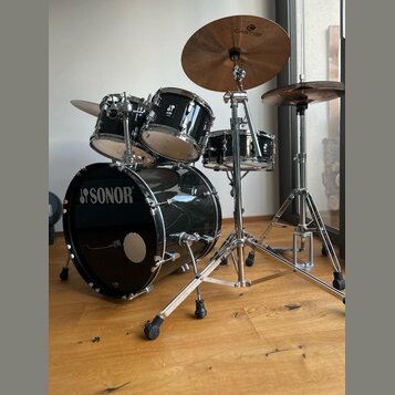 Černá bicí souprava značky SONOR s chromovaným kováním. Sestává z basového bubnu, dvou tomů a malého bubnu, k nimž jsou připojeny dva činely, z nichž…