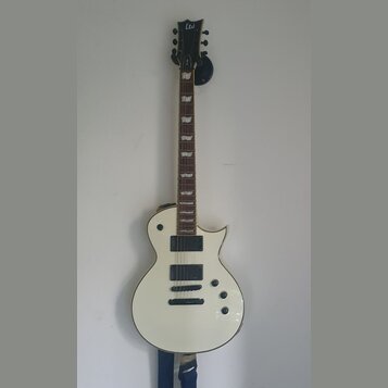 Bílá elektrická kytara s černým hmatníkem a dvěma humbuckery. Na hlavě je bílé logo Ltd. Kytara má čtyři ovládací knoflíky a je umístěna na nástěnním…