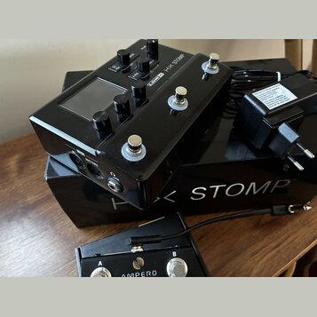 Černý efektový pedál s nápisem LINE 6 HX STOMP, vybavený displejem, několika ovládacími knoflíky a třemi stoupakovými spínači. Nástroj leží na černé…