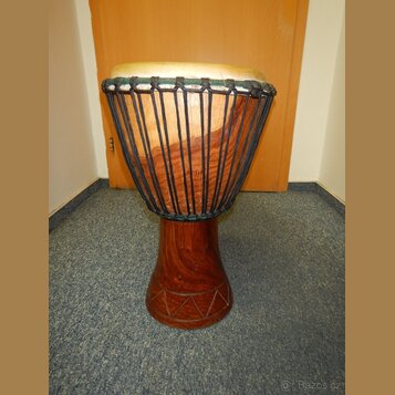Dřevěný buben typu djembe s hnědým povrchem a světlou koženou membránou. Tělo nástroje je v horní části opletené černým šňůrem a v dolní části má vyř…