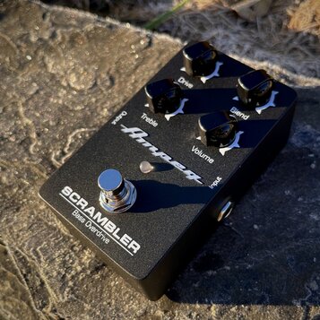 Černý kytarový pedál s nápisem SCRAMBLER Bass Overdrive a logem Ampeg. Na horní straně jsou čtyři černé otočné knoflíky s popisky Drive, Blend, Trebl…