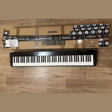 Černý digitální klavír s bílými a černými klávesami. Na horní hraně je nápis CASIO, označení Privia a model PX-S3100 BK. Nástroj leží na dřevěné podl…