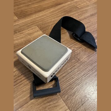 Dřevěný cajon v podobě malého kostky s šedým povrchem na horní straně. Na bočních stěnách jsou černé nápisy „WOODY“ a „TELL WOOD“ s motivem stromu. N…