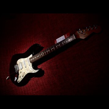 Černá elektrická kytara typu Stratocaster s bílým perleťovým pickguardem. Na hlavě hředu je viditelné logo Fender. Kytara má tmavý hmatník a na krku…
