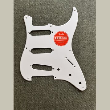 Bílá pickguard pro elektrickou kytaru s výřezy pro tři snímače. Na povrchu je nalepená červená kulatá nálepka s nápisem Fender FREE ONLINE LESSONS FO…