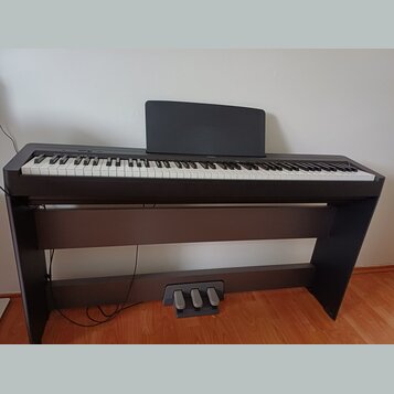 Digitální piano v černé barvě s bílými a černými klávesami, umístěné na matchingovém černém stojanu. Součástí je černý notový pouzdro a pedálová jedn…