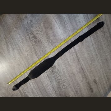 Černý popruh na hudební nástroj z textilního materiálu s širší středovou částí. Popruh je položen vedle žlutého skladovacího metru na šedé podlaze. N…