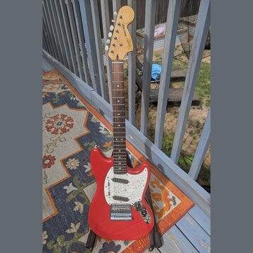 Elektrická kytara s červeným trupem a bílým pickguardem. Na hlavě hředu je nápis "Squier" a modelové označení "MUSTANG". Hmatník je tmavého dřeva, ky…