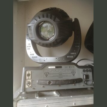 Černý světelný efekt typu moving head s kovovou konstrukcí a čočkou v přední části. Pod ním je šedý ovládací panel s konektory, přepínačem a úchytkou…