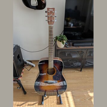 Akustická kytara s tělem v provedení sunburst (černá, hnědá a žlutá). Na hlavě je bílý nápis YAMAHA. Nástroj stojí na černém stojanu v interiéru.