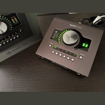 Šedý zvukový interface Apollo Twin značky Universal Audio. Zařízení má velký centrální ovládací knoflík, LED indikátory úrovně a řadu tlačítek s nápi…