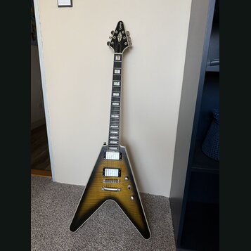 Elektrická kytara s typickým V-tvarem těla v žlutém gradientním provedení s černým okrajem. Na hlavě je viditelné logo Gibson. Kytara má dva humbucke…