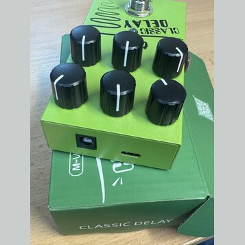 Zelený kytarový efekt s šesti černými oválnými knoflíky. Na horní straně je nápis CLASSIC DELAY a grafický symbol vlnovky. Efekt leží na zelené krabi…