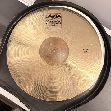 Zlatý činel s koncentrickým žilováním, umístěný v černém obalu. Na povrchu je bílý štítek s nápisy pAiste, Twenty a TRADE MARK MADE IN SWITZERLAND. V…