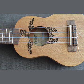 Dřevěný ukulele s vyřezaným otvorem v těle, který je zdoben vypáleným motivem želvy. Uvnitř otvoru je vidět štítek s nápisy Keiki, Ukuleles a čárový…