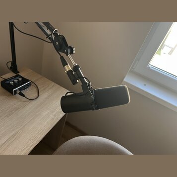 Na fotografii je černý mikrofon s nasazenou šedou větrolnicí, který je upevněn na nastavitelné rameni. Na těle mikrofonu je viditelný symbol CE a neč…
