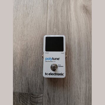 Bílý kytarový ladič s černým displejem. Na přední straně jsou nápisy „display“, „pitch“, „tuning“, „output“ a „input“. Uprostřed je modrý a černý náp…