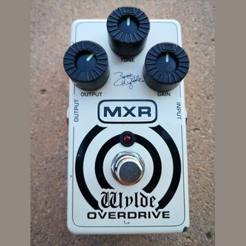 Bílý kytarový efekt pedál s nápisy MXR a Wylde OVERDRIVE. Obsahuje tři černé ovládací knoflíky s popisky OUTPUT, TONE a GAIN. Na šasi je viditelný po…
