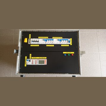 Elektrický rozvaděč v kovovém kufru s černým dnem. Horní část obsahuje jističe a štítky s nápisy PHASE, Fi SCHALTER-OUT1, Fi SCHALTER-OUT2, LED LICHT…