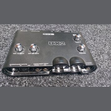 Černý zvukový interface Line 6 UX2. Na horní straně jsou čtyři chromované otočné ovladače s nápisy MIC 1/L GAIN, MIC 2/R GAIN, OUTPUT a PHONES. V pře…